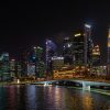 Night view of a lit city skyline with a curved pedestrian bridge over calm water, at night in Marina Bay (Singapore).