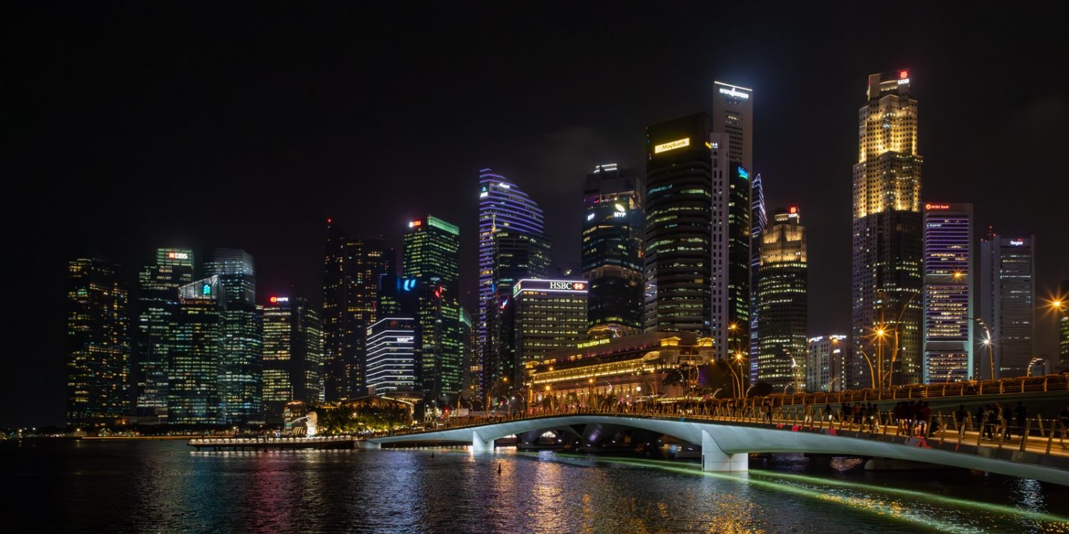 Night view of a lit city skyline with a curved pedestrian bridge over calm water, at night in Marina Bay (Singapore).