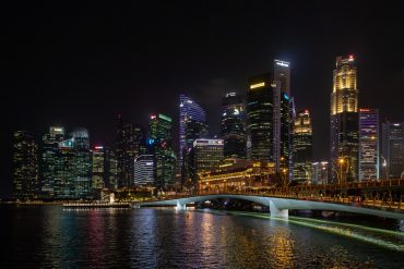 Night view of a lit city skyline with a curved pedestrian bridge over calm water, at night in Marina Bay (Singapore).