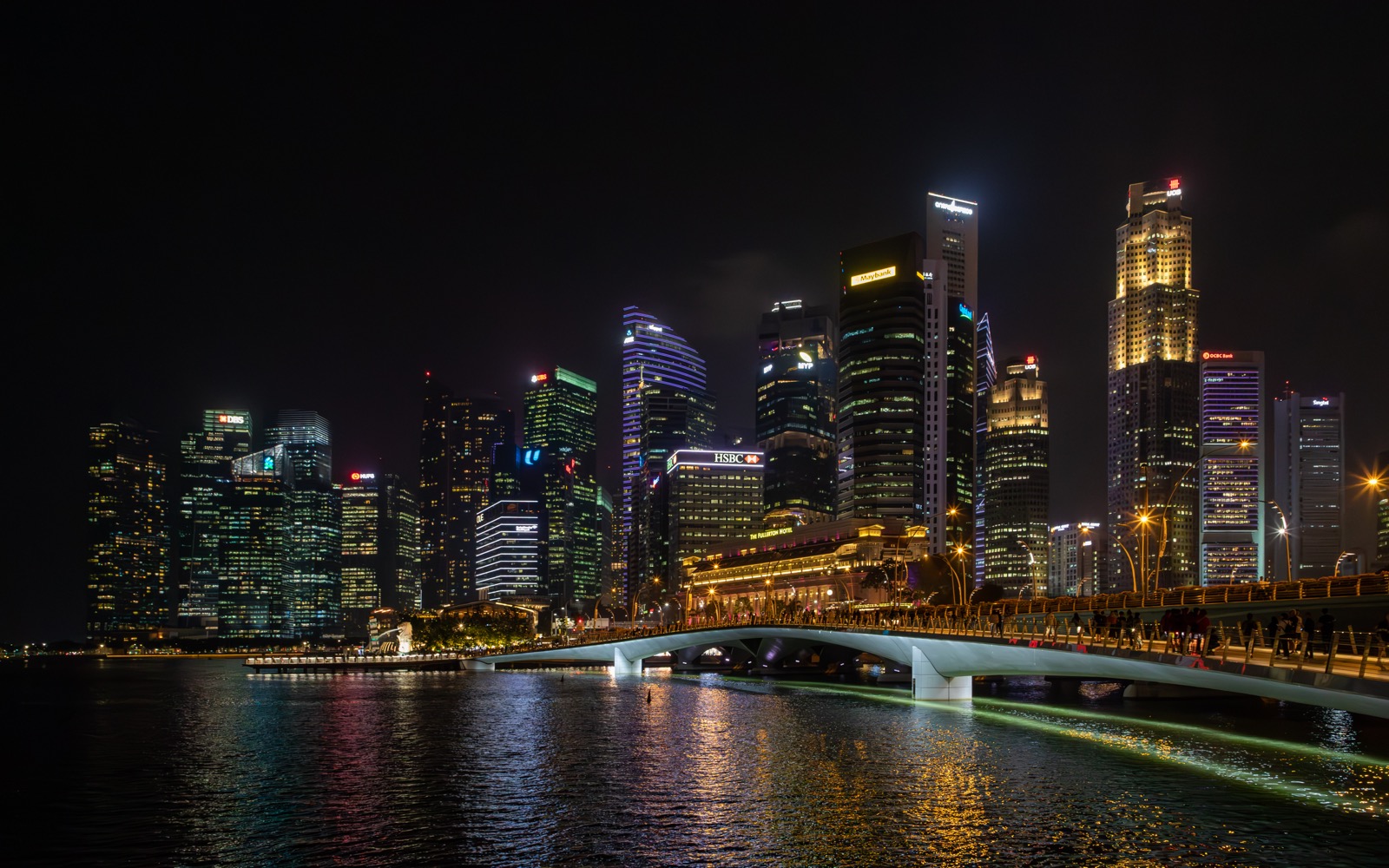 Night view of a lit city skyline with a curved pedestrian bridge over calm water, at night in Marina Bay (Singapore).