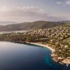 Aerial view of a hillside resort town along a crescent beach, with white buildings, green trees, and turquoise bay water. - Possibly a luxury hotel complex on a coastal hillside.
