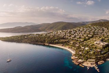 Aerial view of a hillside resort town along a crescent beach, with white buildings, green trees, and turquoise bay water. - Possibly a luxury hotel complex on a coastal hillside.