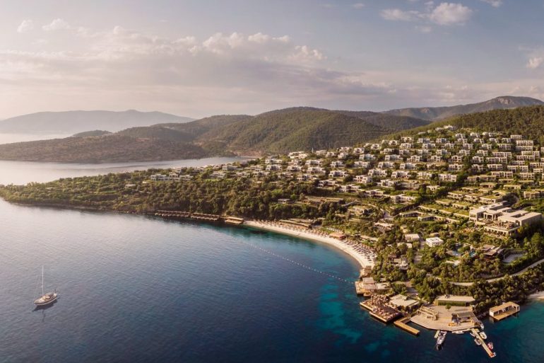 Aerial view of a hillside resort town along a crescent beach, with white buildings, green trees, and turquoise bay water. - Possibly a luxury hotel complex on a coastal hillside.