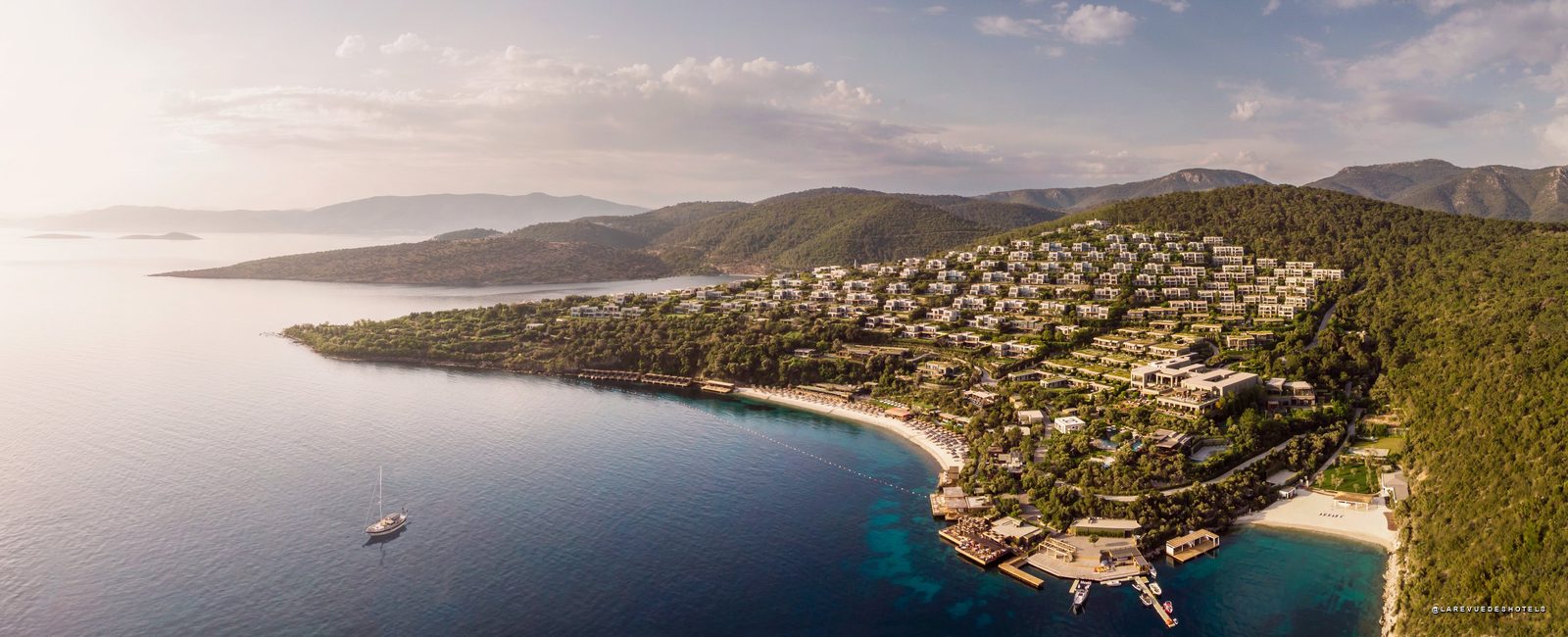 Aerial view of a hillside resort town along a crescent beach, with white buildings, green trees, and turquoise bay water. - Possibly a luxury hotel complex on a coastal hillside.