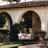 A woman lounges on a cushioned outdoor sofa under an arched patio, reading a book beside flowering pots.
