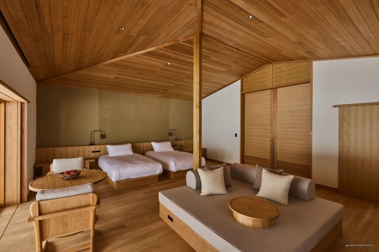 Wood-clad hotel suite with two low platform beds, a sofa area, and a round coffee table in warm lighting