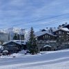 Snowy alpine village with chalet-style buildings, a Christmas tree, and a gondola lift against snow-capped mountains.