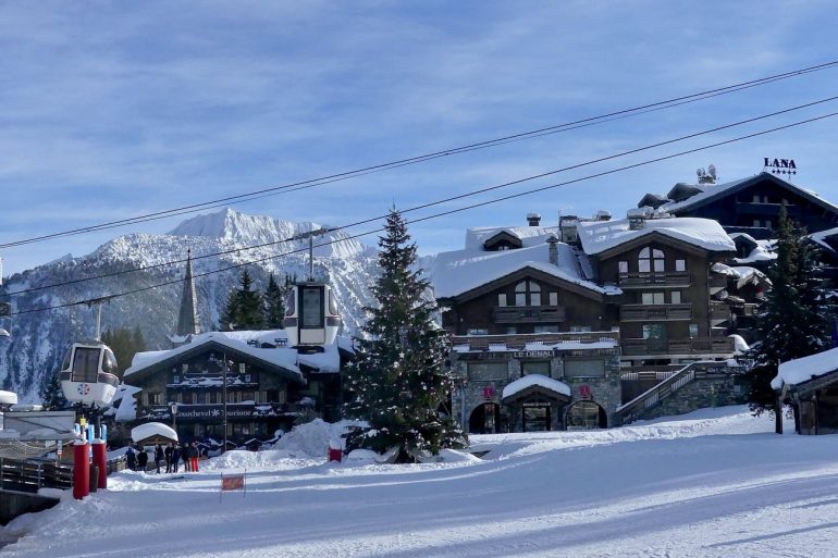 Snowy alpine village with chalet-style buildings, a Christmas tree, and a gondola lift against snow-capped mountains.
