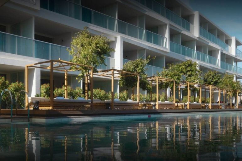 Modern hotel with glass balconies and a wooden poolside cabana lounge along a reflective pool in front of the building.