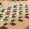 Rows of turquoise beach umbrellas with matching lounge chairs on a sandy shore, creating a grid pattern and long shadows from the sun.