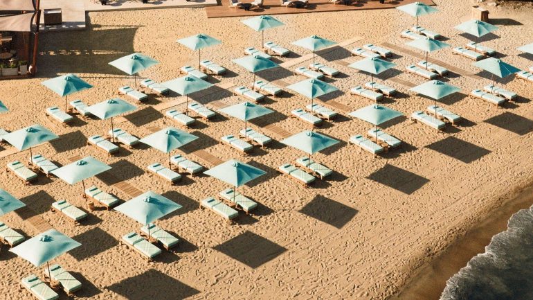 Rows of turquoise beach umbrellas with matching lounge chairs on a sandy shore, creating a grid pattern and long shadows from the sun.