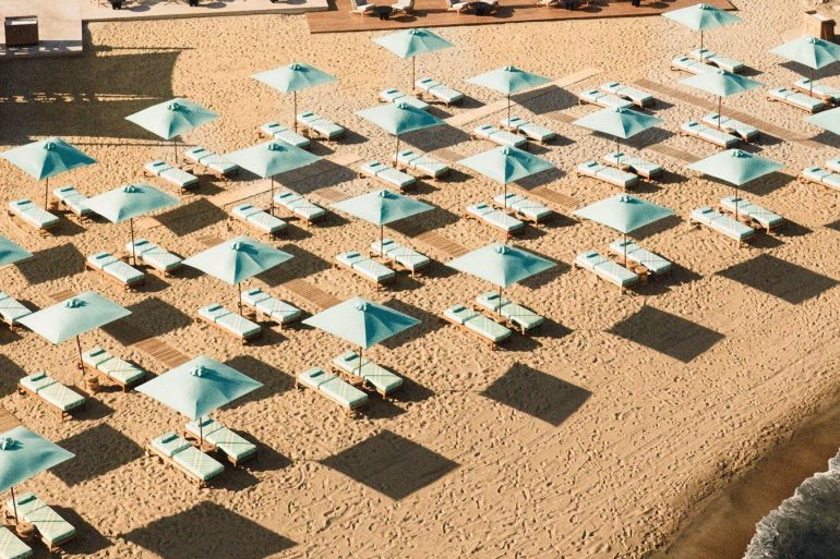 Rows of turquoise beach umbrellas with matching lounge chairs on a sandy shore, creating a grid pattern and long shadows from the sun.