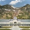 Grand neoclassical building with a central domed pavilion, set against forested mountains under a blue sky.