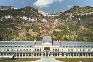 Grand neoclassical building with a central domed pavilion, set against forested mountains under a blue sky.