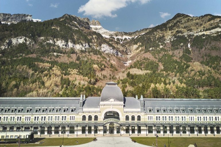 Grand neoclassical building with a central domed pavilion, set against forested mountains under a blue sky.