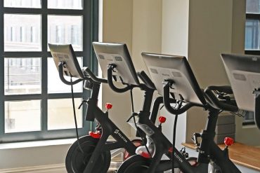 Row of Peloton stationary bikes in a bright spin studio by a large arched window, screens up front.