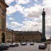 Historic city square with beige neoclassical buildings, a tall column monument at center, and cobblestone streets with cars and pedestrians nearby.