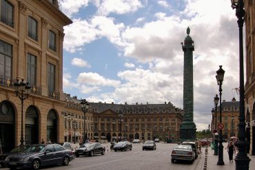 Historic city square with beige neoclassical buildings, a tall column monument at center, and cobblestone streets with cars and pedestrians nearby.