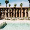 Resort hotel with beige multi-story balconies lined by palm trees, pool in the foreground and sun loungers nearby.