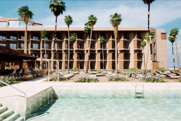 Resort hotel with beige multi-story balconies lined by palm trees, pool in the foreground and sun loungers nearby.