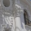White ornate classical facade with decorative columns and a small balcony with a wrought-iron railing against a blue sky.