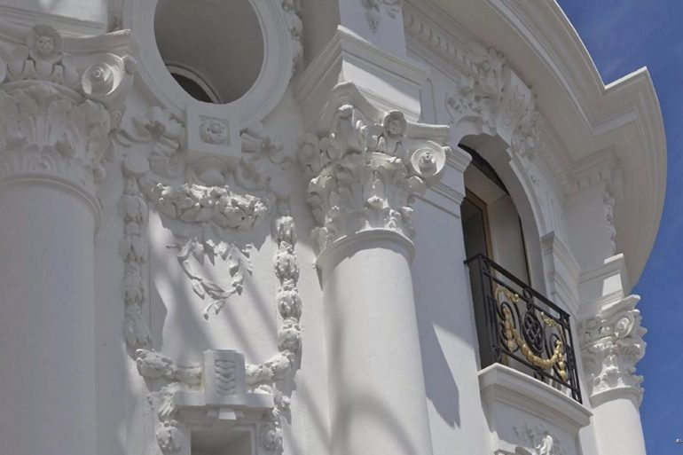 White ornate classical facade with decorative columns and a small balcony with a wrought-iron railing against a blue sky.