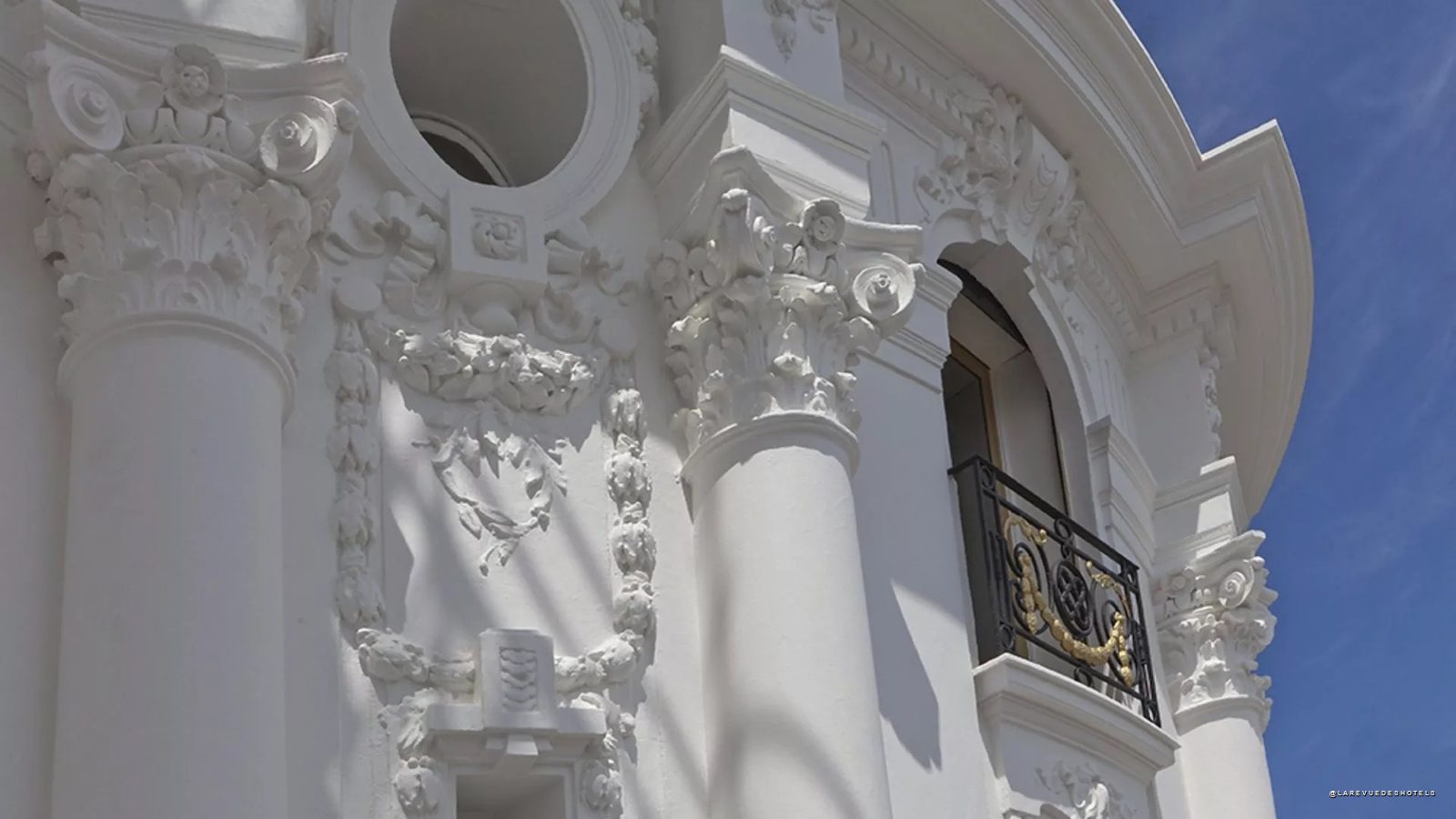White ornate classical facade with decorative columns and a small balcony with a wrought-iron railing against a blue sky.
