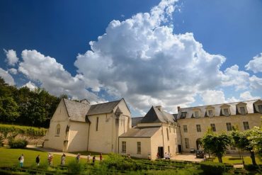 Historic stone building with pitched roofs and arched windows, set in a formal garden on a sunny day with a blue sky and large clouds.