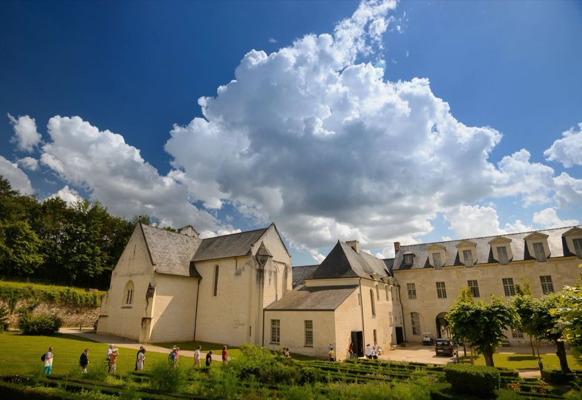 Historic stone building with pitched roofs and arched windows, set in a formal garden on a sunny day with a blue sky and large clouds.
