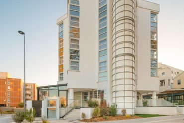 Modern white building with a curved glass tower and tall vertical windows on a street corner at sunset.