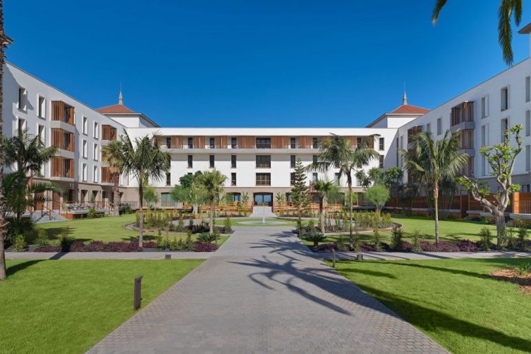 Hotel courtyard with palm trees, green lawn, and white buildings surrounding a central paved walkway.