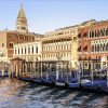 Gondolas tied to posts along a Venetian canal with ornate historic buildings in the background, reflections on the water. The scene includes a tall clocktower and red-brick facades typical of Venice.