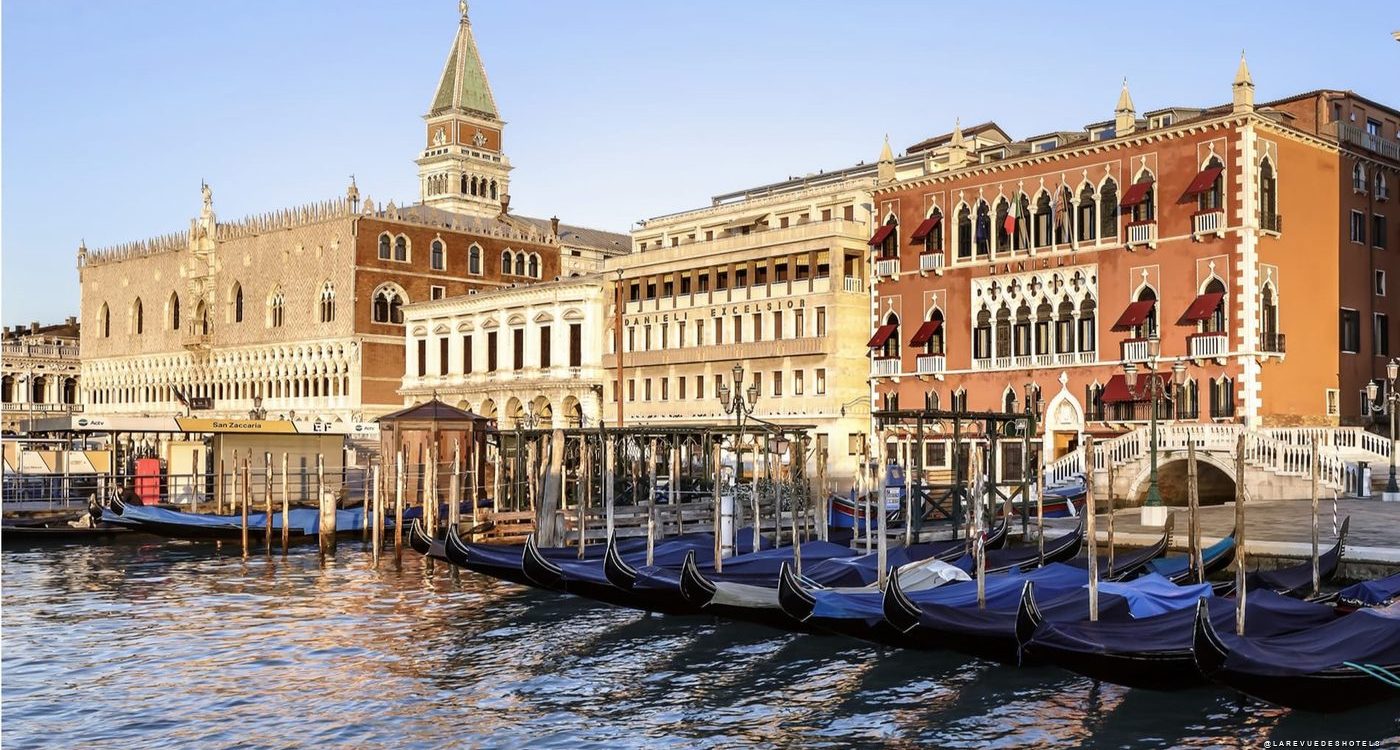 Gondolas tied to posts along a Venetian canal with ornate historic buildings in the background, reflections on the water. The scene includes a tall clocktower and red-brick facades typical of Venice.