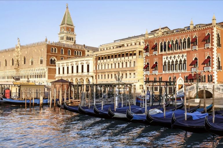 Gondolas tied to posts along a Venetian canal with ornate historic buildings in the background, reflections on the water. The scene includes a tall clocktower and red-brick facades typical of Venice.