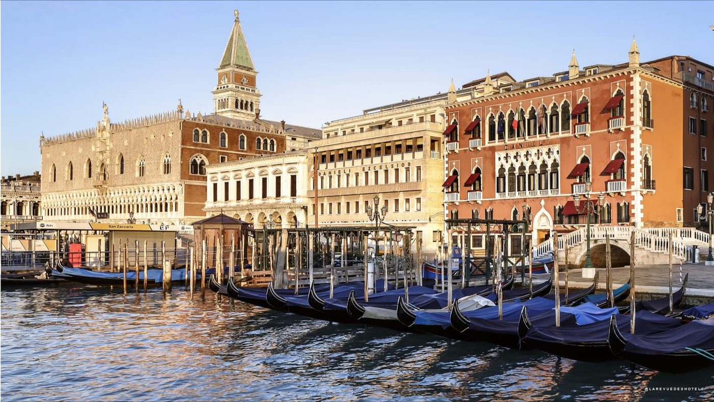 Gondolas tied to posts along a Venetian canal with ornate historic buildings in the background, reflections on the water. The scene includes a tall clocktower and red-brick facades typical of Venice.