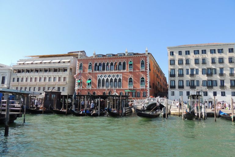 Gondolas docked along a Venetian canal with ornate historic buildings and a blue sky above.