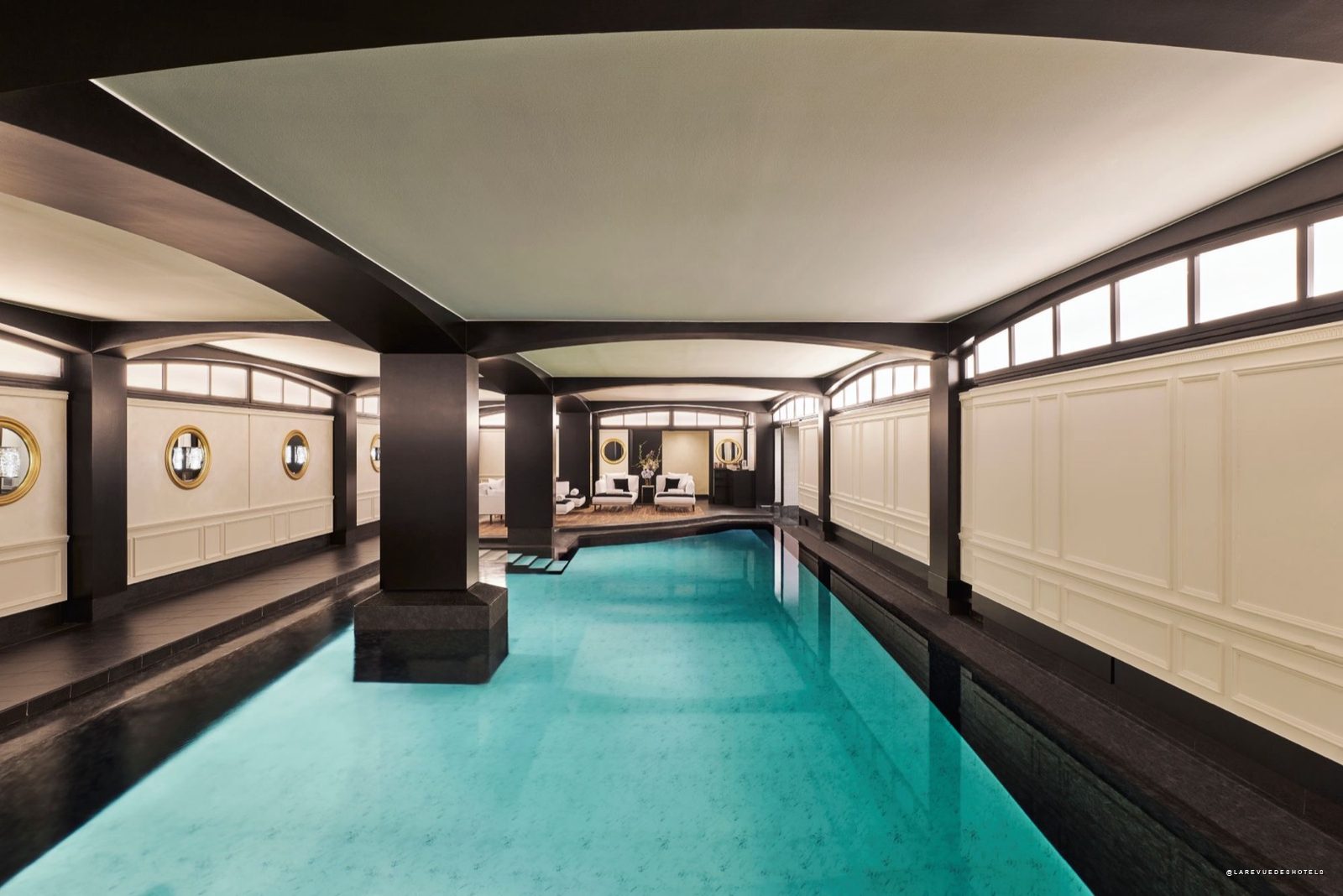 Indoor luxury hotel pool with black pillars and white-paneled walls, seating area visible at far end.