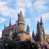 Stone Hogwarts-style castle with tall spires perched on a rocky hill, framed by trees under a blue, partly cloudy sky.