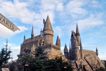 Stone Hogwarts-style castle with tall spires perched on a rocky hill, framed by trees under a blue, partly cloudy sky.
