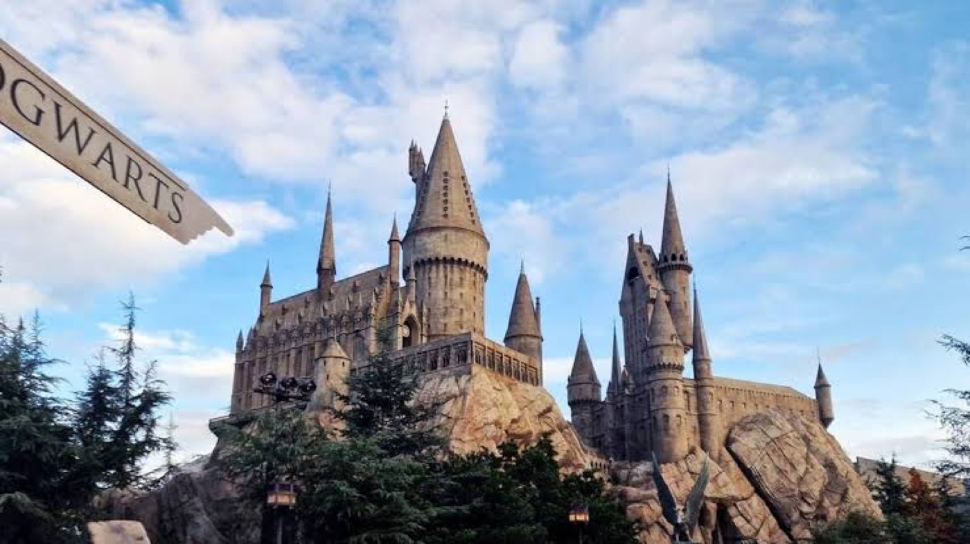 Stone Hogwarts-style castle with tall spires perched on a rocky hill, framed by trees under a blue, partly cloudy sky.