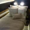 Queen bed in a modern hotel room with white pillows and beige bedding, illuminated by wall lamps above.”] ,