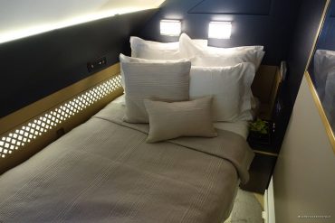 Queen bed in a modern hotel room with white pillows and beige bedding, illuminated by wall lamps above.”] ,