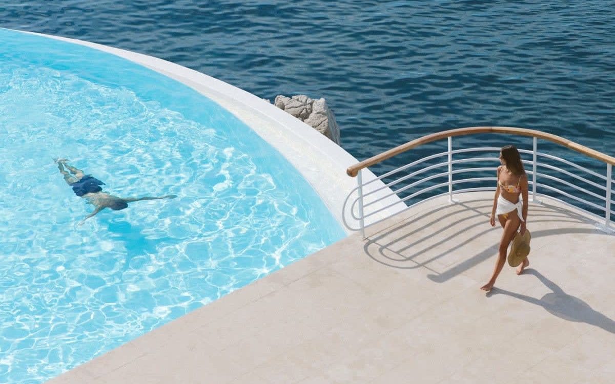 Woman in a bikini walking on a pool deck overlooking the sea, carrying a hat.