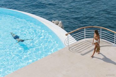 Woman in a bikini walking on a pool deck overlooking the sea, carrying a hat.