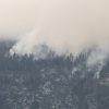 Smoke rising through a mountainous forest as a wildfire burns among the trees, with a pale sky above.