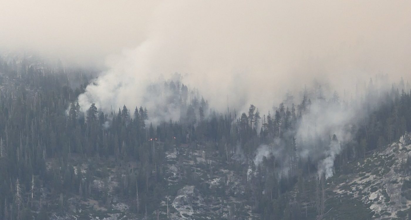 Smoke rising through a mountainous forest as a wildfire burns among the trees, with a pale sky above.