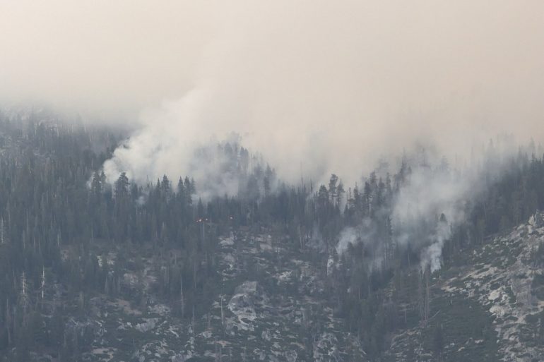 Smoke rising through a mountainous forest as a wildfire burns among the trees, with a pale sky above.