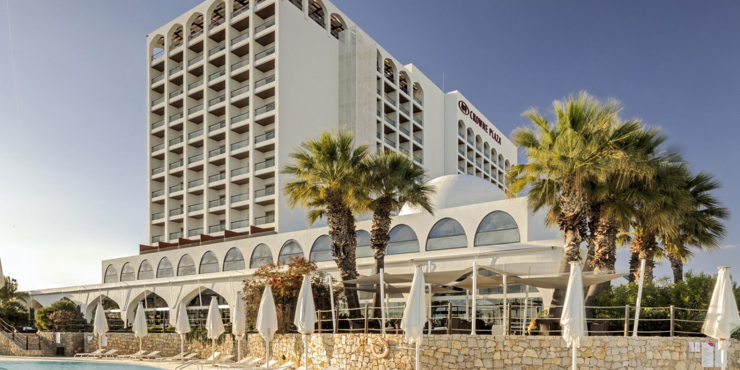 Exterior view of Crowne Plaza hotel with a pool area, white arched architecture, and palm trees in the foreground.
