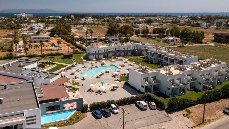 Aerial view of a seaside resort with white apartments, a central pool area, and sun loungers around the pool.