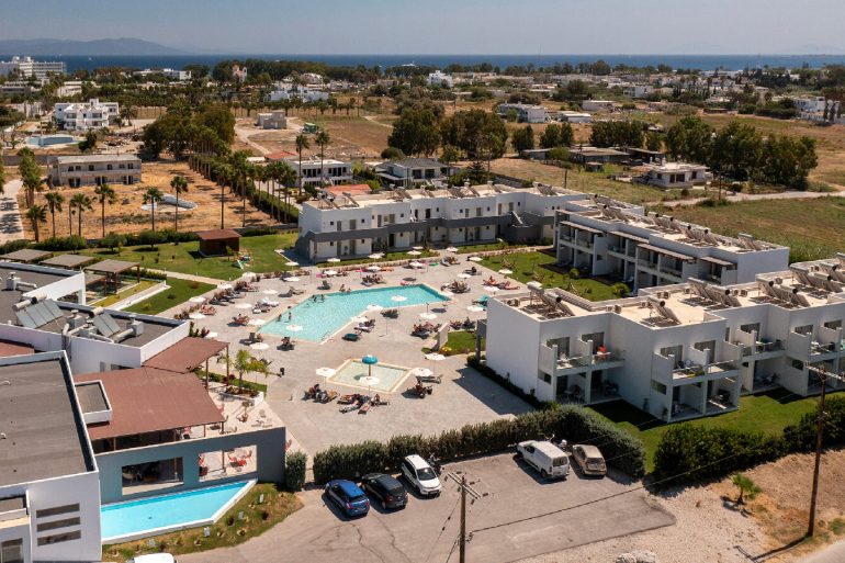 Aerial view of a seaside resort with white apartments, a central pool area, and sun loungers around the pool.
